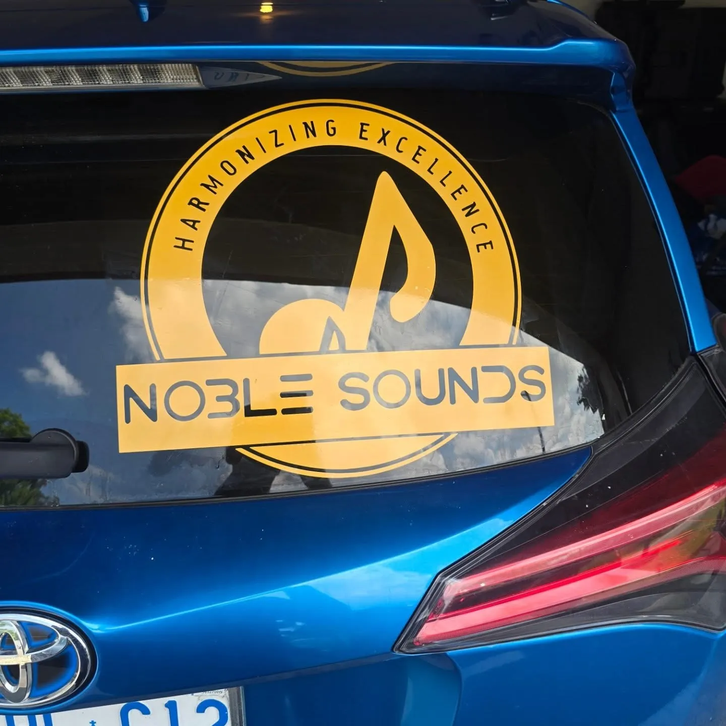 Yellow Noble Sounds roundel and wordmark applied to the rear window of a blue hatchback