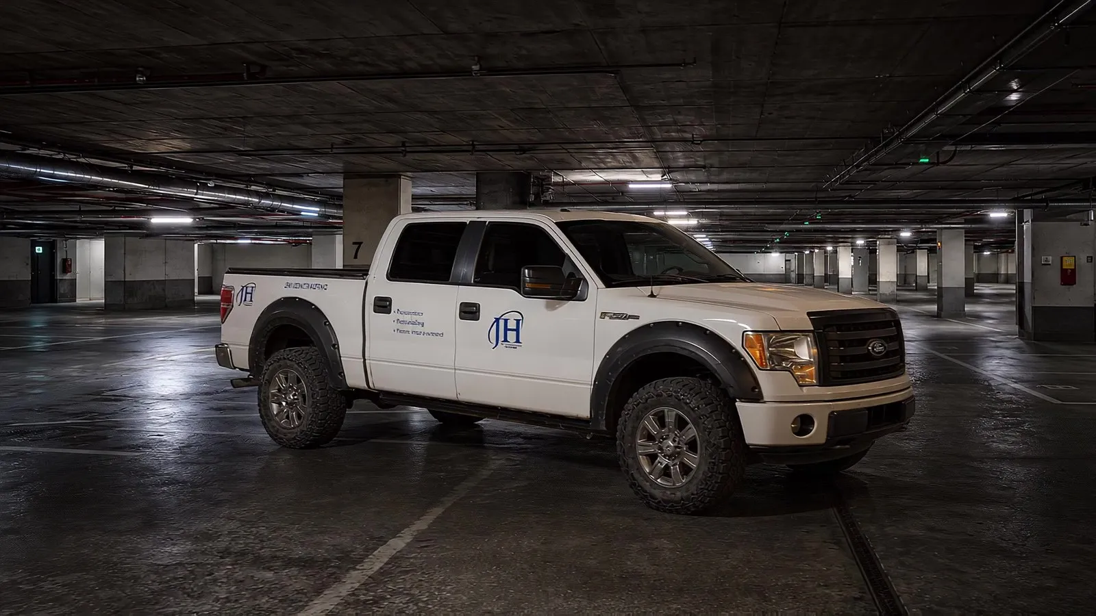 White crew-cab pickup carrying blue JH Contracting door decals, parked under a garage light